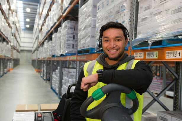 Smiling worker in a bright warehouse, wearing a neon safety vest and headset, leans on a cart. Shelves filled with neatly stacked boxes line the aisle.