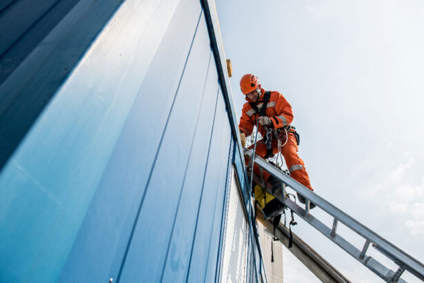 Worker in orange safety gear on a ladder against a blue building, adjusting ropes, under a clear sky. Scene conveys professionalism and focus.