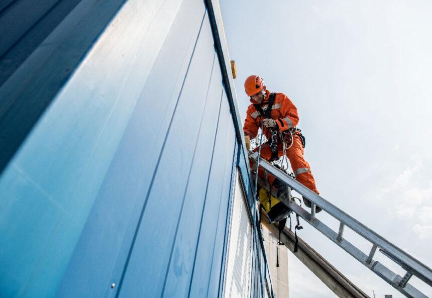 Worker in orange safety gear on a ladder against a blue building, adjusting ropes, under a clear sky. Scene conveys professionalism and focus.