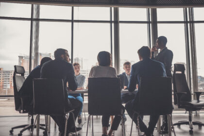 A diverse group of people sits around a conference table in a modern office, with cityscape views through large windows, conveying a professional tone.