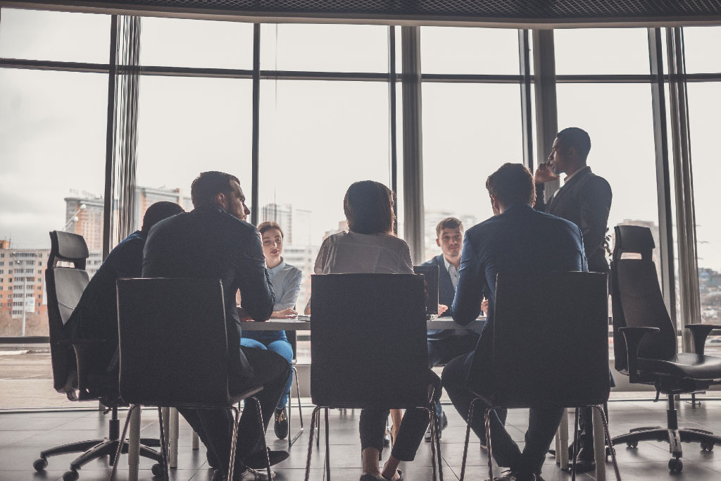 A diverse group of people sits around a conference table in a modern office, with cityscape views through large windows, conveying a professional tone.