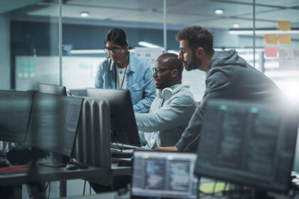 Three people work intently at a computer in a modern office. Monitors display code, suggesting a tech or programming environment. Collaborative atmosphere.