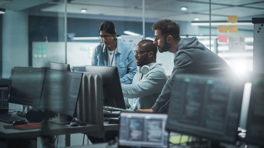 Three people work intently at a computer in a modern office. Monitors display code, suggesting a tech or programming environment. Collaborative atmosphere.
