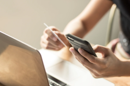 A person uses a smartphone with one hand and holds a credit card in the other, beside an open laptop, suggesting online shopping or banking.