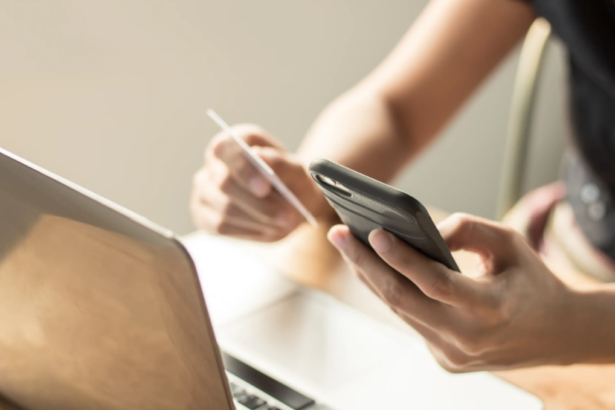 A person uses a smartphone with one hand and holds a credit card in the other, beside an open laptop, suggesting online shopping or banking.
