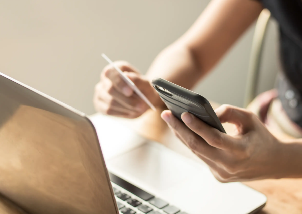 A person uses a smartphone with one hand and holds a credit card in the other, beside an open laptop, suggesting online shopping or banking.