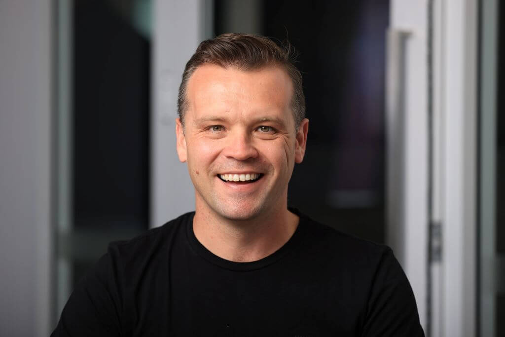 A man with short hair, wearing a black shirt, smiles warmly while sitting indoors. Soft lighting and a blurred background create a friendly atmosphere.