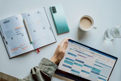 Overhead view of a tidy workspace featuring an open weekly planner with handwritten notes, a pen, a smartphone, a tablet displaying a digital calendar, a cup of coffee and a glass of water, illustrating the use of both paper and digital tools for scheduling and time management.