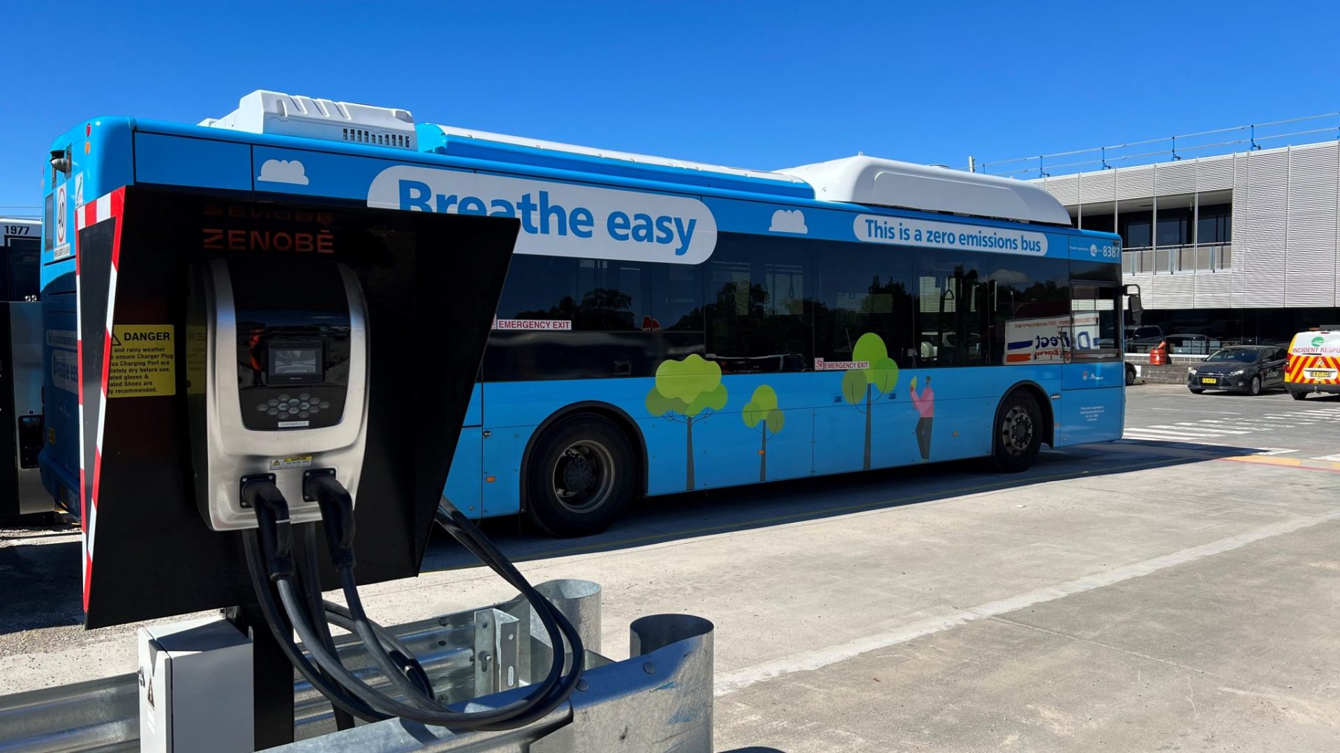 A blue zero-emissions bus is parked beside a Zenobē electric charging unit, with charging cables connected in a depot setting.