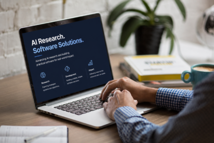 A person types on a laptop displaying an "AI Research. Software Solutions." webpage featuring sections on research, development and impact. The laptop sits on a desk beside a notebook and coffee cup, with a plant and books blurred in the background.