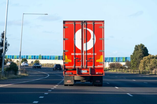 Rear view of an Australia Post delivery truck travelling along a multi-lane road, with the company's red branding visible on the trailer and light traffic ahead under a clear blue sky.