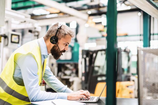A warehouse worker wearing a high-visibility safety vest and headset uses a laptop at a workstation inside an industrial facility, with warehouse equipment and machinery visible in the background.