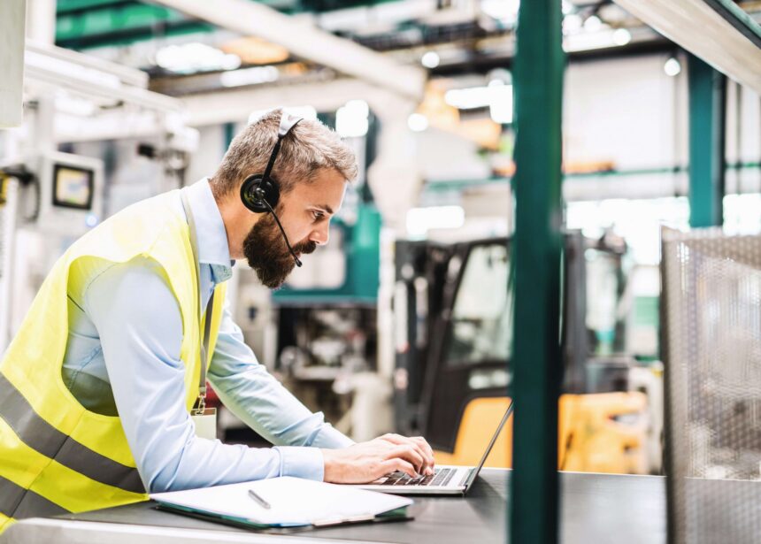 A warehouse worker wearing a high-visibility safety vest and headset uses a laptop at a workstation inside an industrial facility, with warehouse equipment and machinery visible in the background.