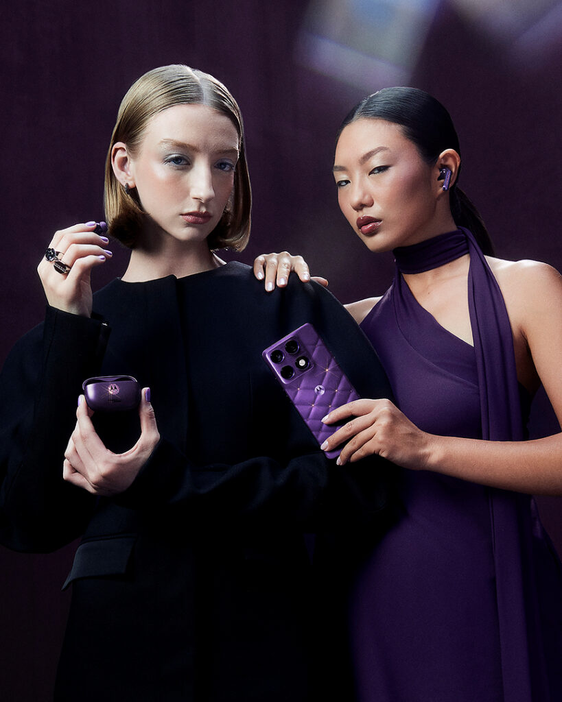 Two models pose against a dark purple background wearing Motorola earbuds, with one holding a purple Motorola smartphone featuring a quilted finish and the other holding a matching purple earbuds charging case.