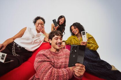 Four people sitting together on a red couch smile and laugh while holding Motorola razr smartphones in different colours, showcasing the devices' foldable design during a group selfie.