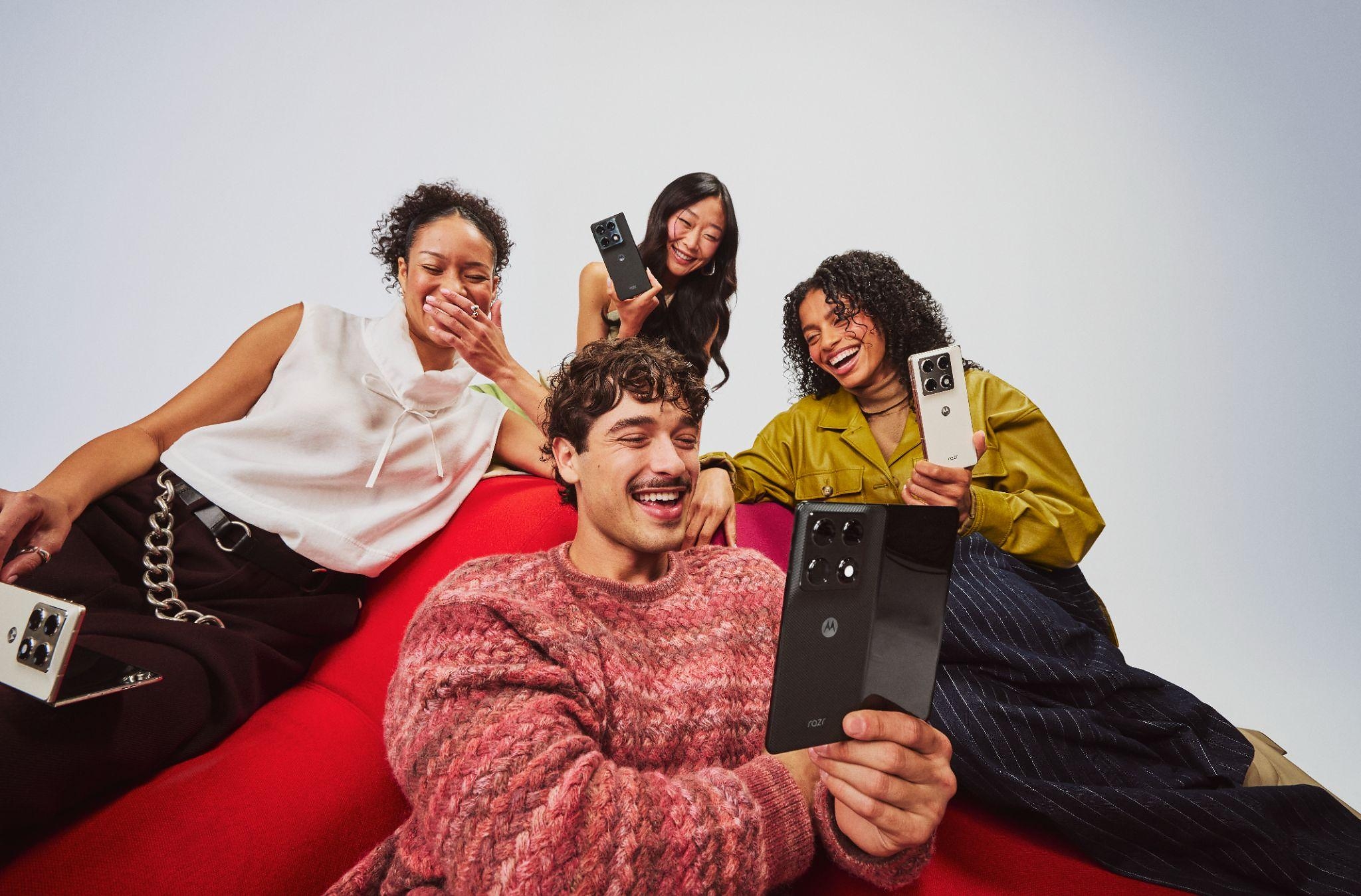 Four people sitting together on a red couch smile and laugh while holding Motorola razr smartphones in different colours, showcasing the devices' foldable design during a group selfie.