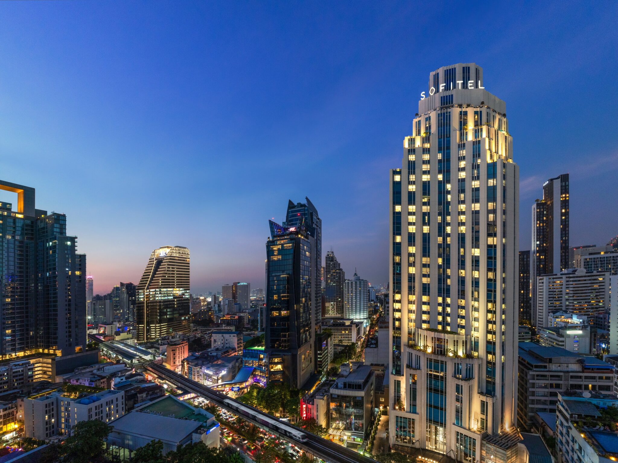 Evening view of Bangkok's skyline featuring the Sofitel Bangkok Sukhumvit hotel illuminated against the cityscape, with surrounding high-rise buildings and the elevated BTS Skytrain line below.
