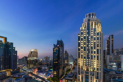 Evening view of Bangkok's skyline featuring the Sofitel Bangkok Sukhumvit hotel illuminated against the cityscape, with surrounding high-rise buildings and the elevated BTS Skytrain line below.