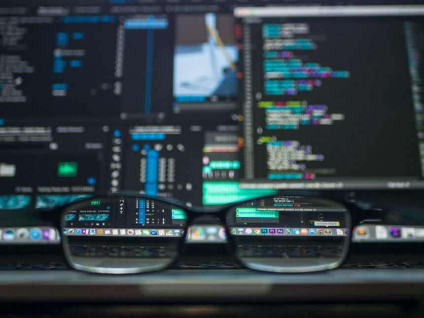 Close-up of a pair of glasses resting on a laptop keyboard, reflecting colourful computer screens displaying code and editing software.