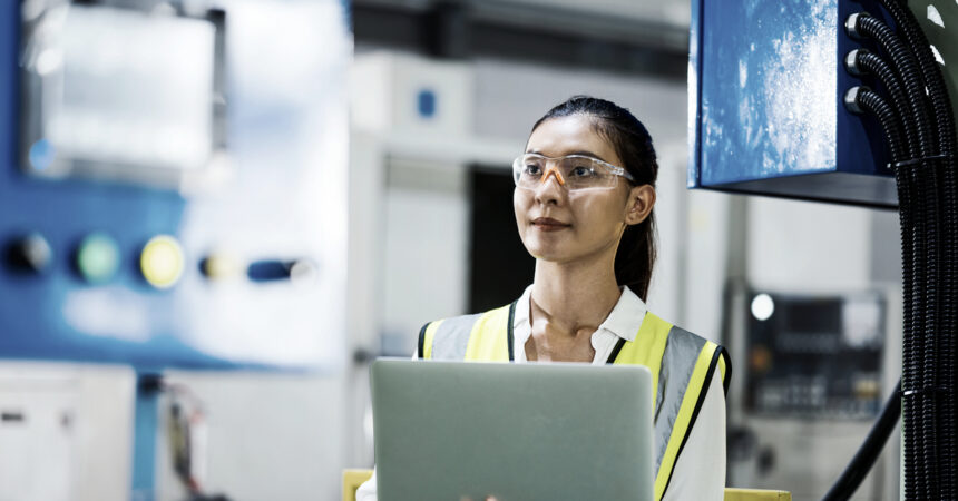 A woman wearing safety glasses holds a laptop in a manufacturing setting.