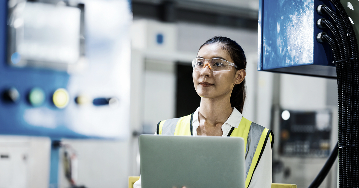A woman wearing safety glasses holds a laptop in a manufacturing setting.