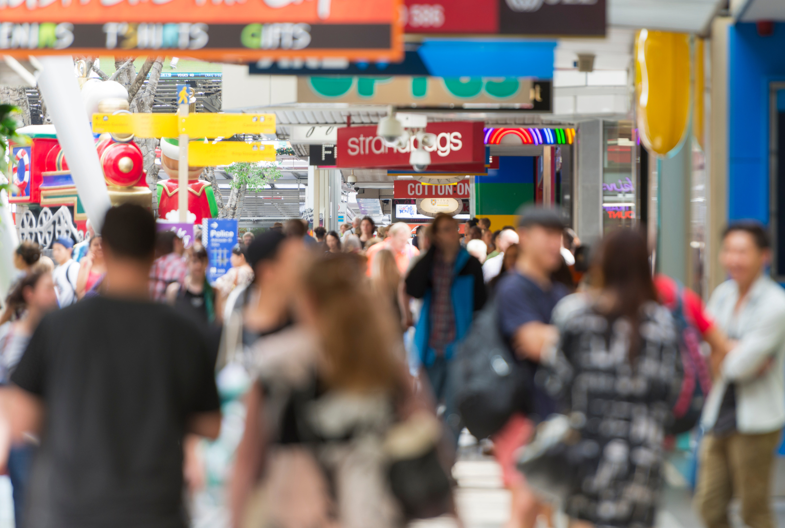 Crowds of shoppers walking through a busy Australian shopping centre, with retail storefronts including Strandbags and Cotton On visible in the background.